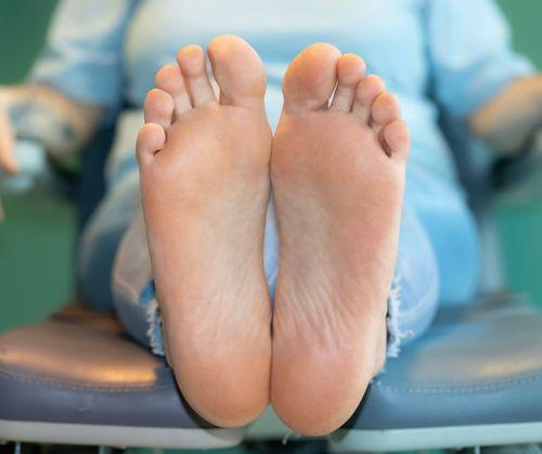 To fødder med bløde såler, der hviler på en stol i en klinik, med en person i baggrunden iført en blå skjorte.