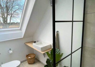 Moderne badeværelse med skråvæg, glasbruser, håndvask på træhylde og grønne planter.