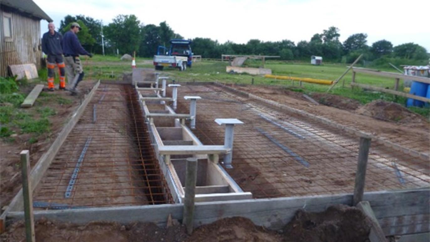 To byggeplads med en betonfundament, hvor arbejdstagere arbejder på at installere støttestrukturer.