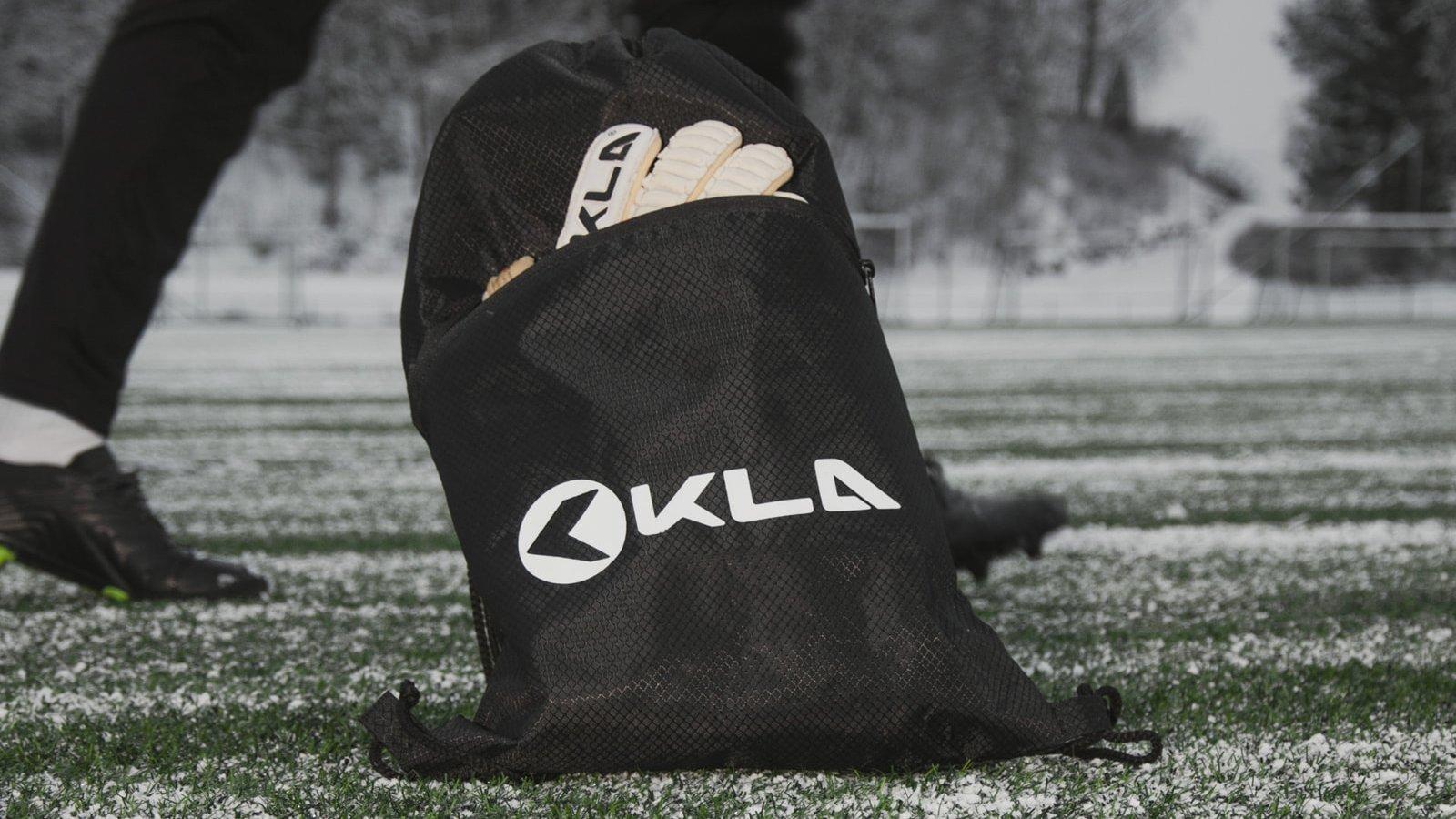 Sort træningspose med KLA-logo og handsker på en fodboldbane dækket af sne.