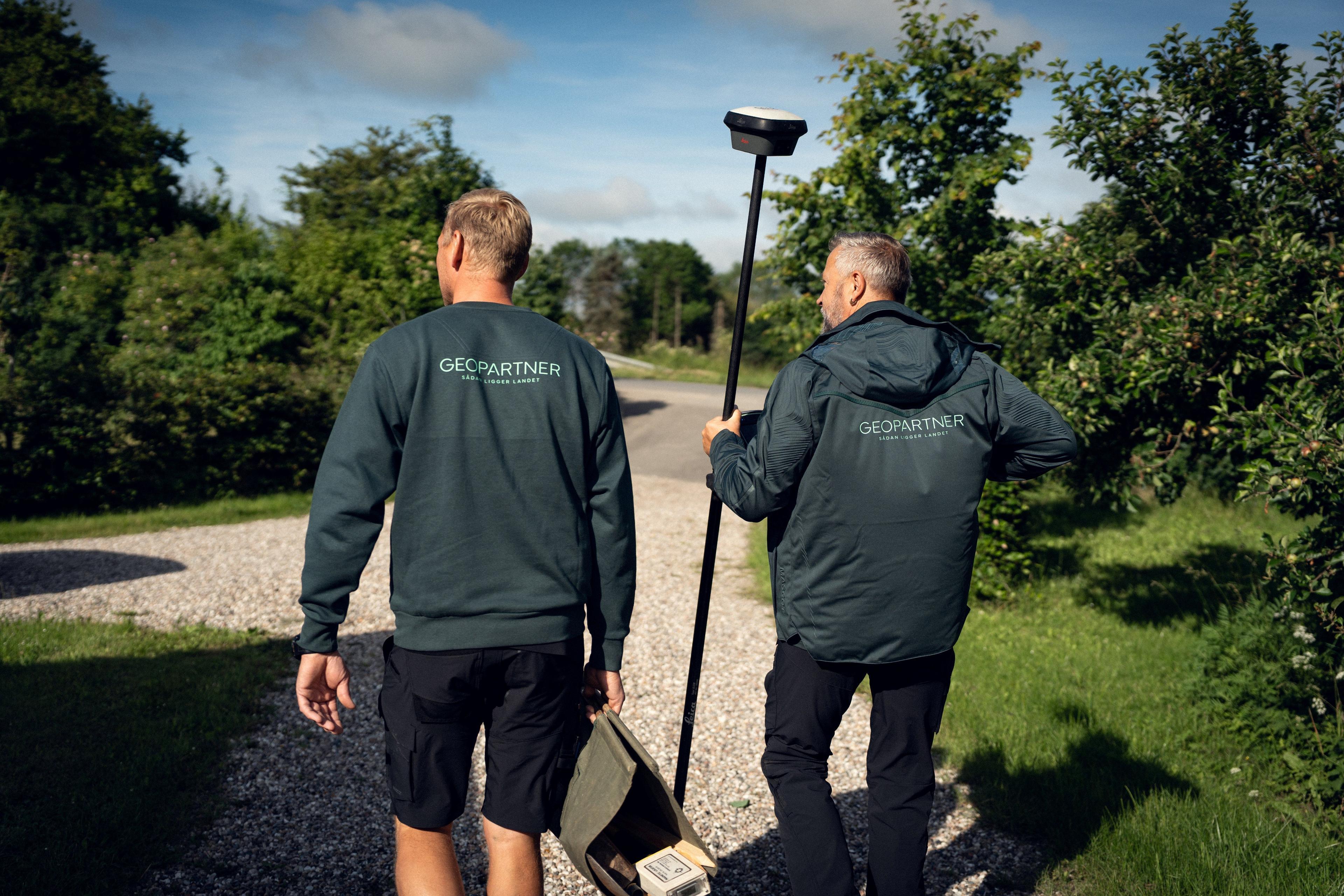 To personer iført mørke jakker går på en grusvej omgivet af grønne træer og buske.