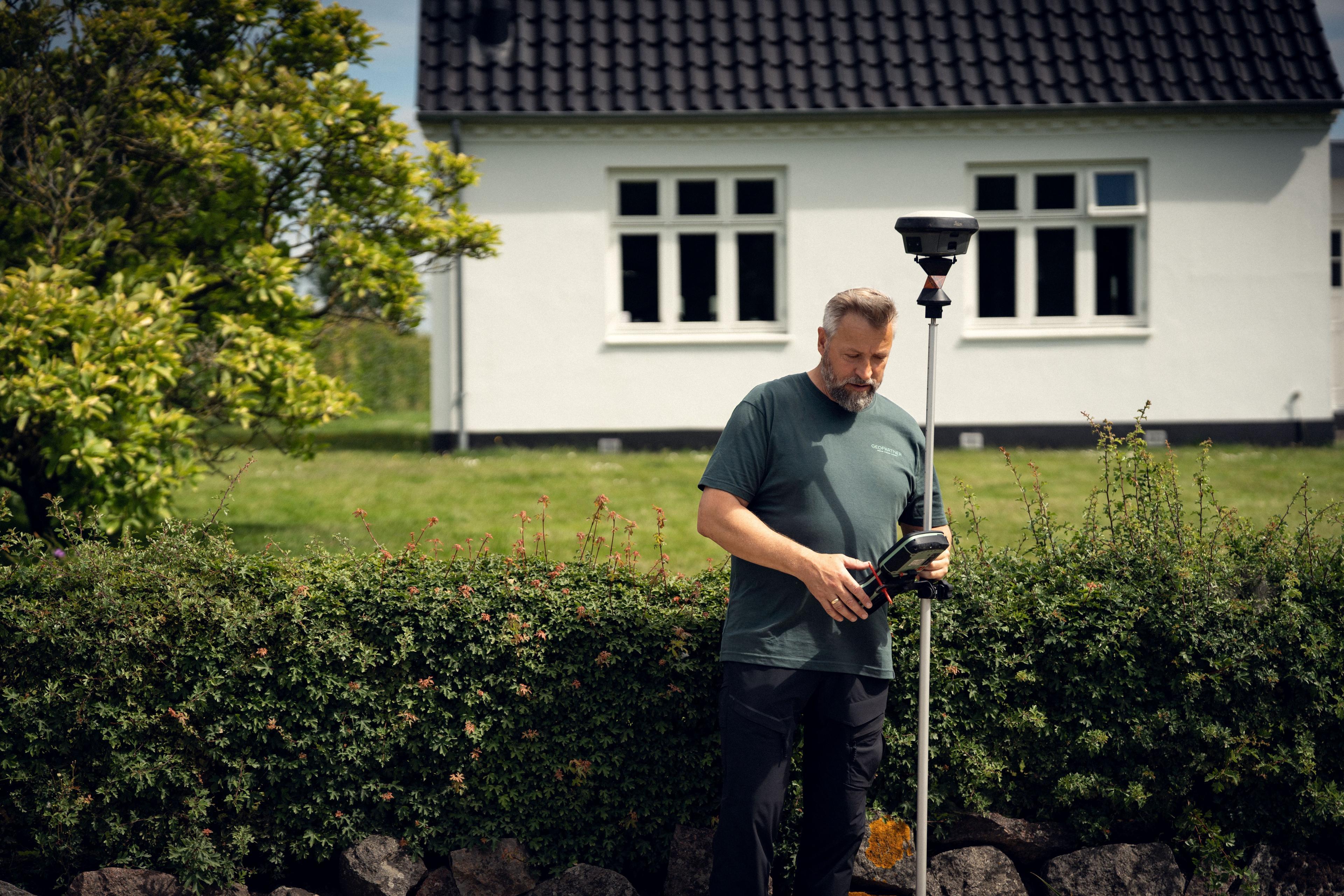 Mand står ved en hæk med en måleapparat i hånden, mens han arbejder på en græsplæne foran et hus.