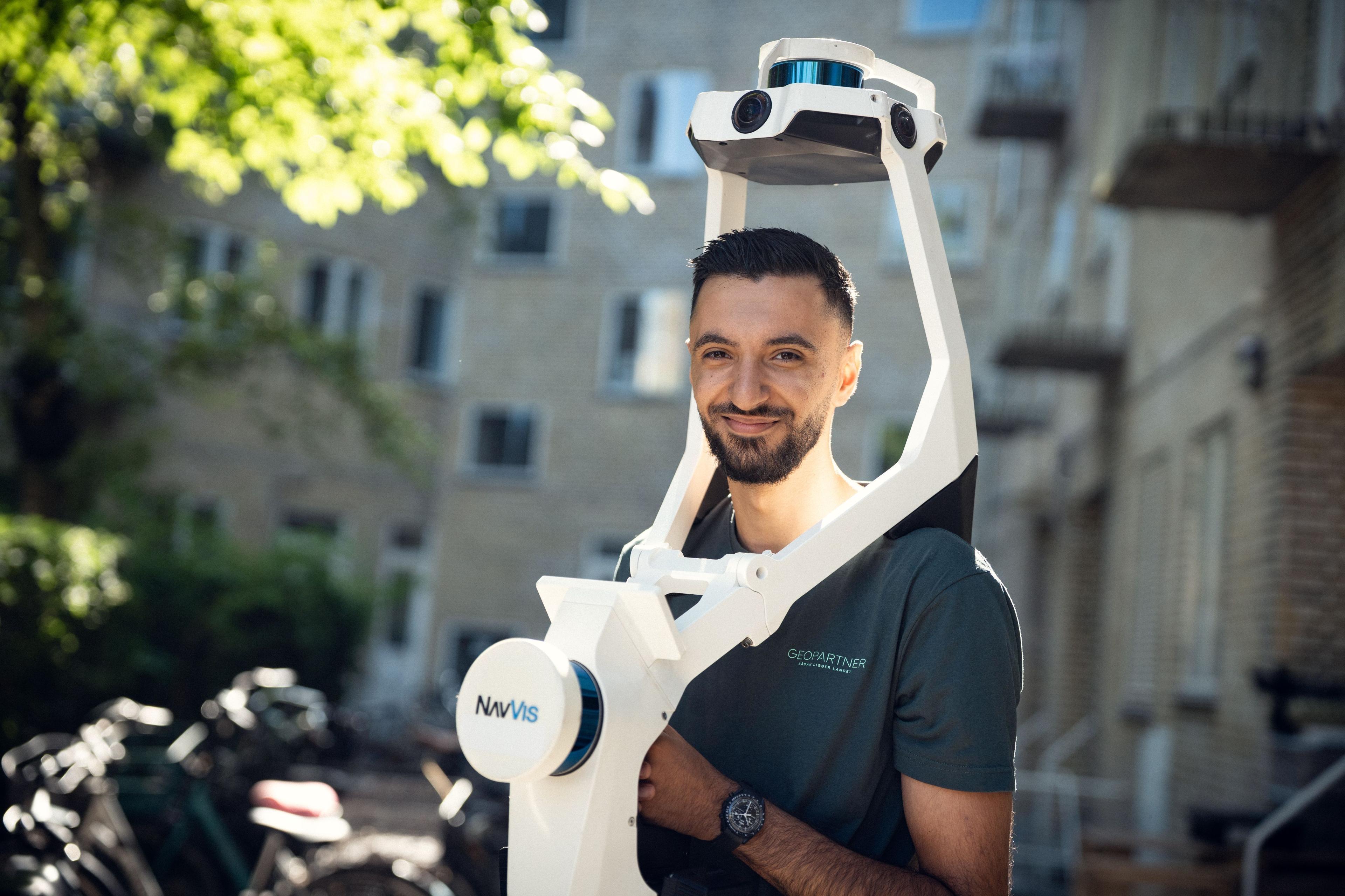 Mand med en avanceret 3D-scanner i hånden, står foran bygninger og cykler i baggrunden.