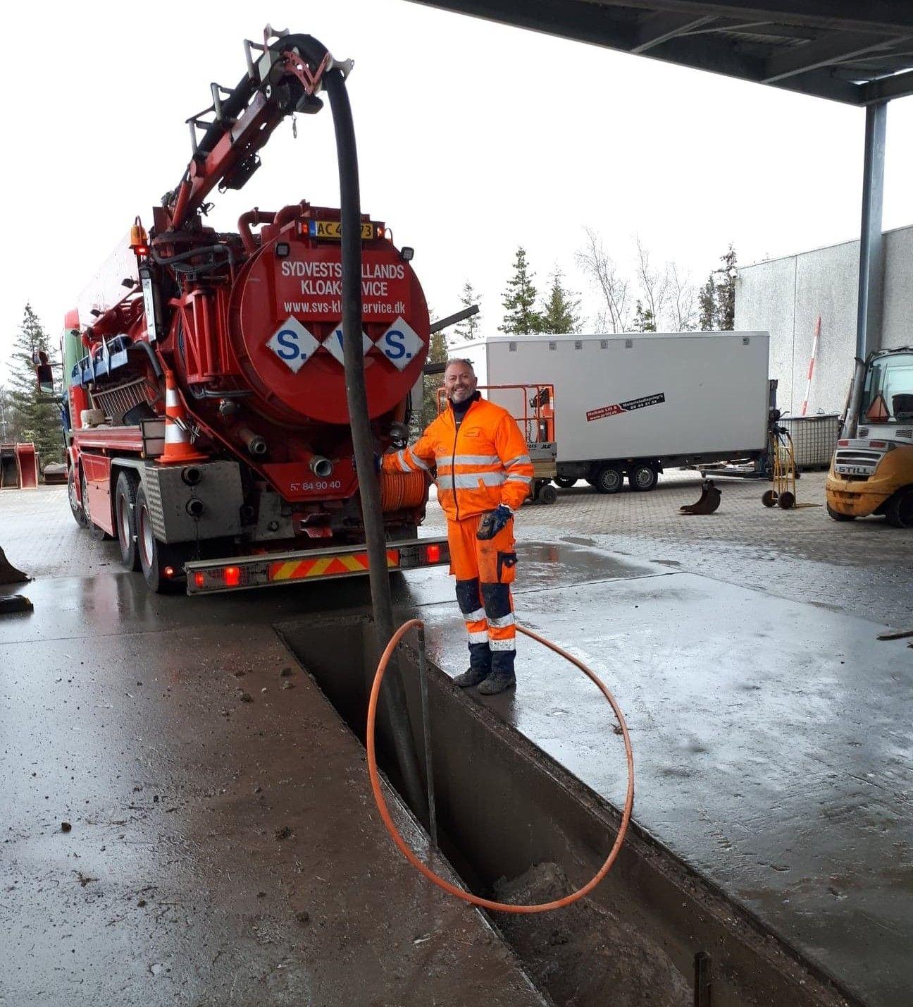 En mand i orange arbejdstøj står ved en stor lastbil med en slange, der er tilsluttet en grav.