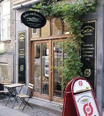 En café med store vinduer, trædøre og grønne planter, beliggende på en gade i København.