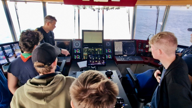 En gruppe unge mennesker ser på skærmene i et skibsførerhus, mens en instruktør viser dem navigationsudstyr.