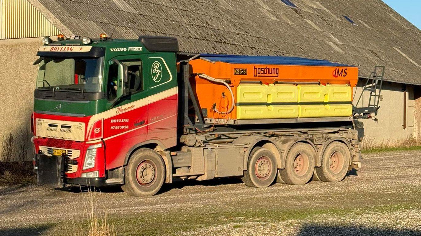 En stor lastbil med en orange og gul kasse kører på en grusvej ved en lade.