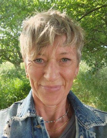 Portræt af en smilende kvinde med kort, lyst hår iført en denimjakke, stående i en grøn have.