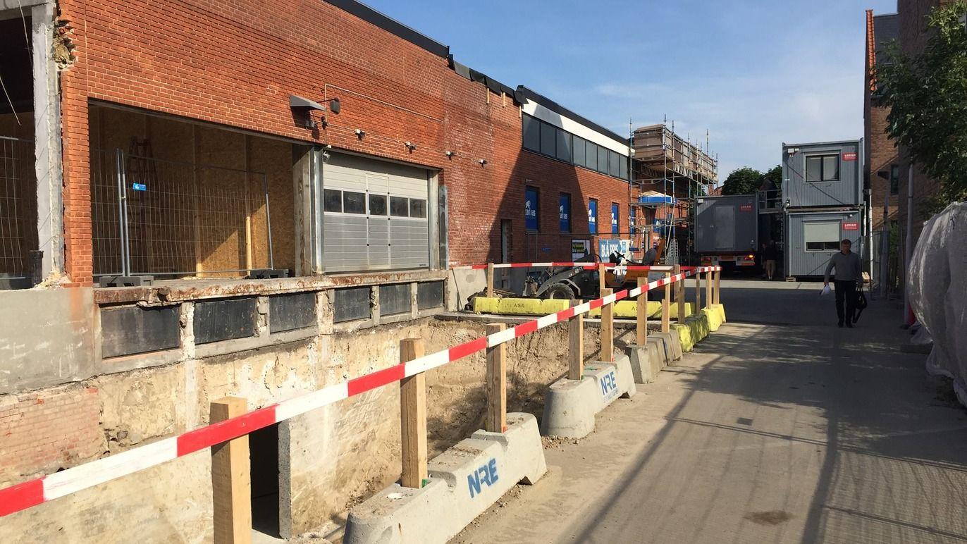 Byggeplads med afskærmning, murstenbygning og gravested til fundamentarbejde under en blå himmel.