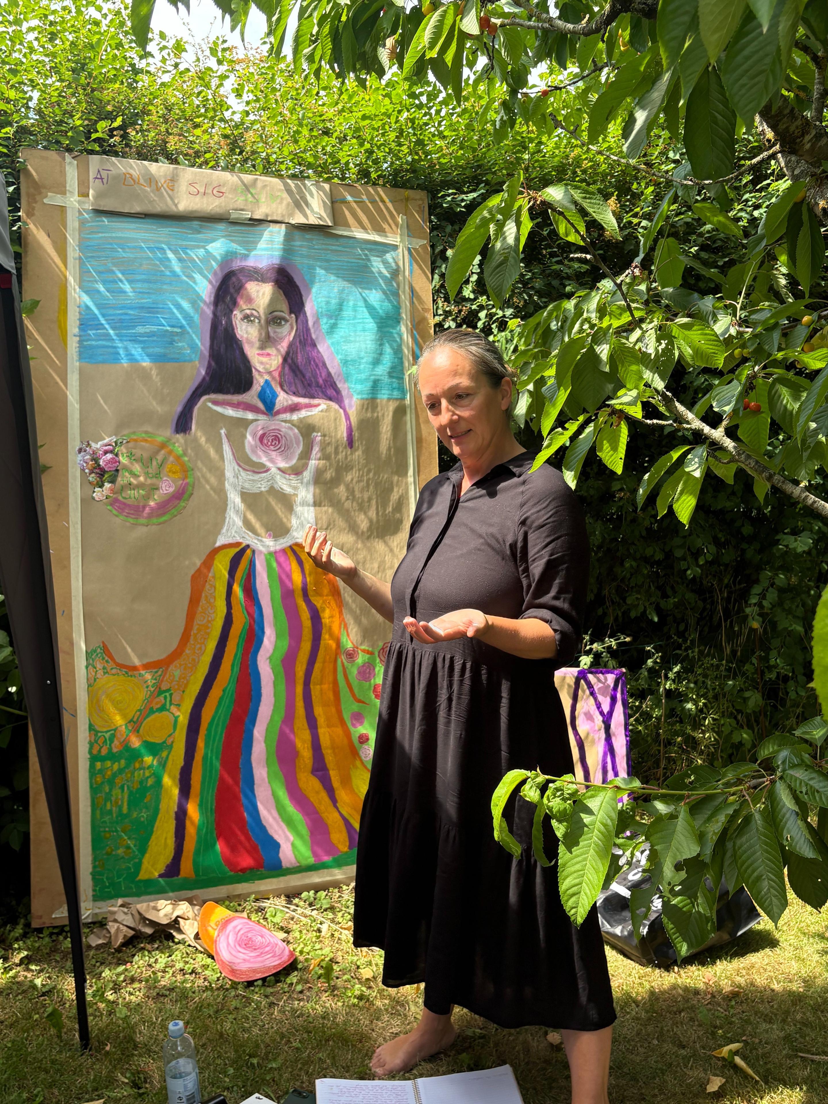 Kunstner i sort kjole præsenterer sit maleri af en farverig figur i haven, omgivet af grønne planter.