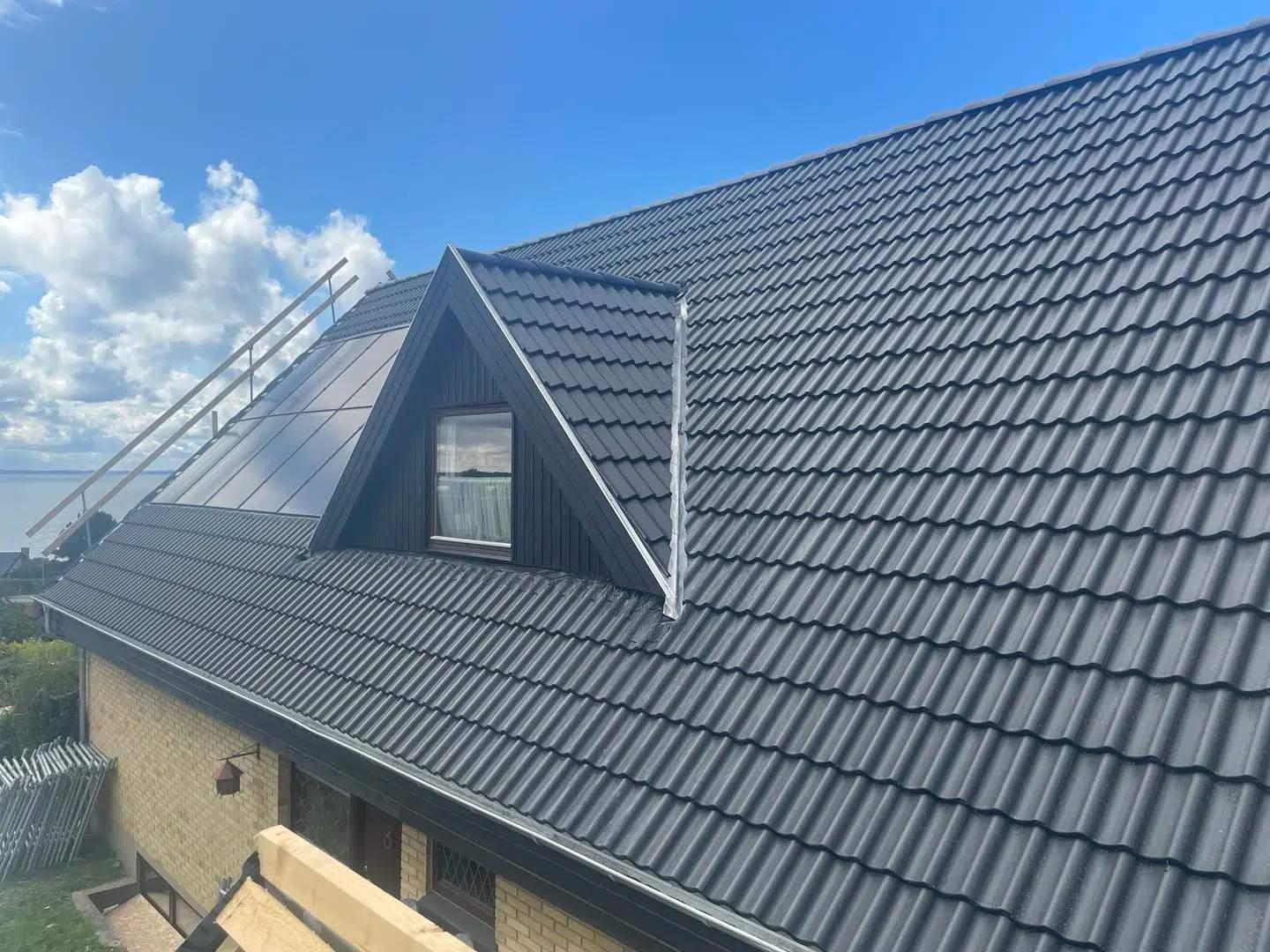 Taget på et hus med sorte tagsten og solpaneler under en blå himmel med skyer.