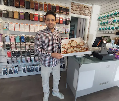 En mand står i en butik med mobiltilbehør og holder en kage i en æske. Bag ham er hylder med telefoncovers.