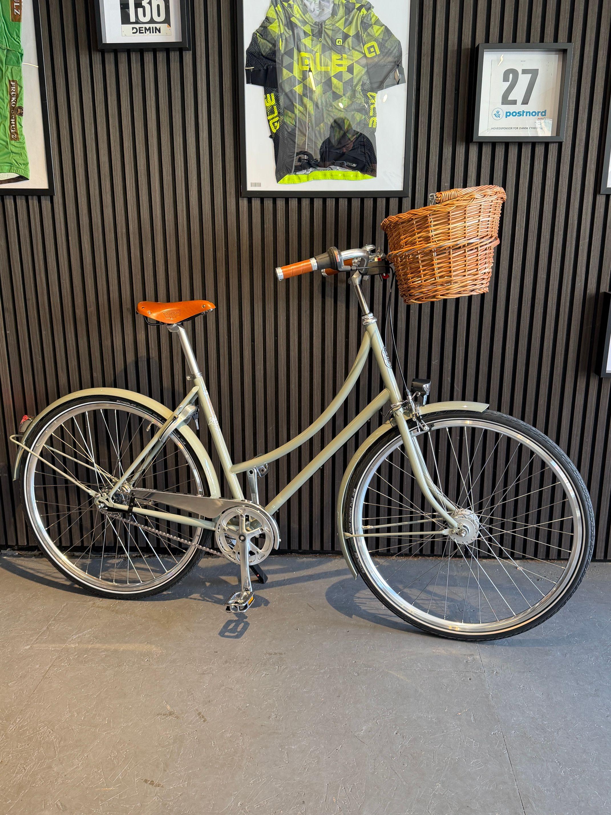 En cykel med kurv og læderbetræk står op ad en væg med billeder i baggrunden.