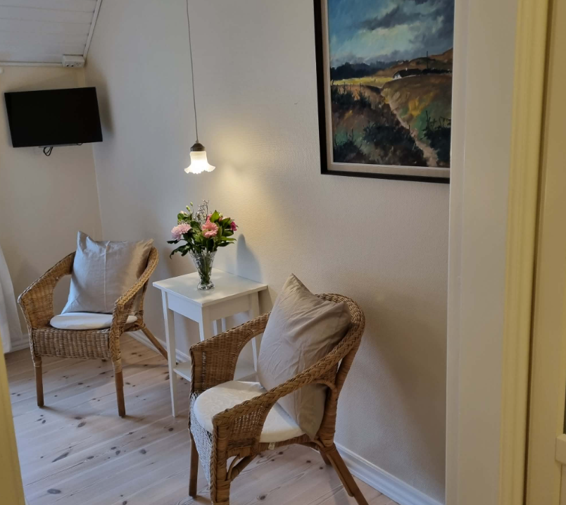 To værelser med kurvestole, et lille bord med blomster og et maleri på væggen i en lys stue.