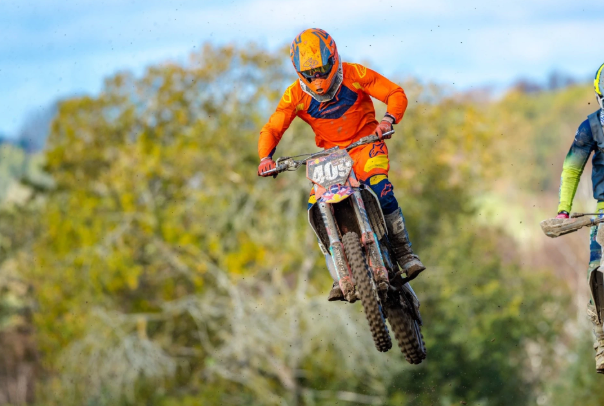 Motocrosskører i orange tøj springer over en forhindring på en grusbane med træer i baggrunden.