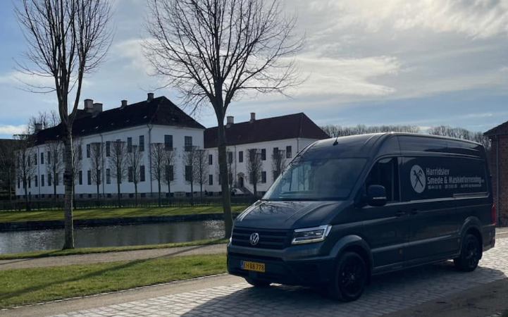 En grå varevogn parkeret ved en kanal med træer og bygninger i baggrunden.