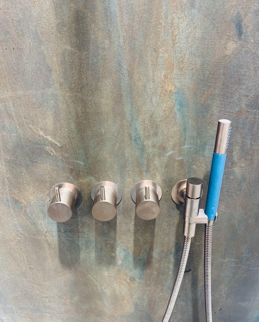 Brusebad med tre metalhåndtag og en håndbruser med blå detalje på en farvet væg.