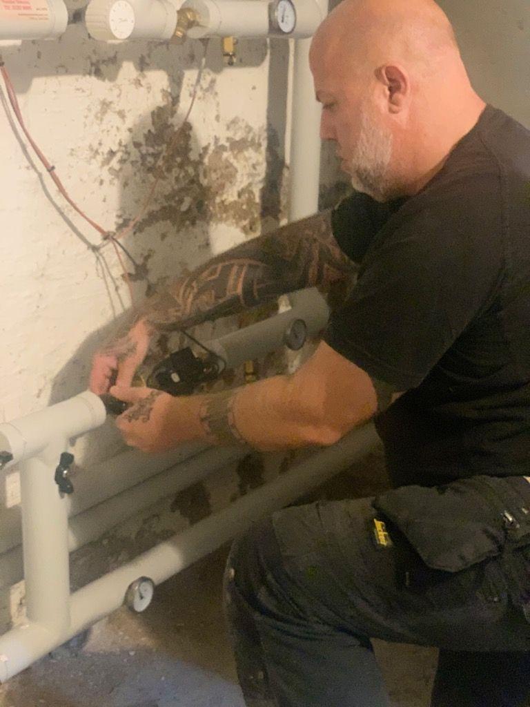 En håndværker arbejder med rørinstallation i en kælder, omgivet af murværk og rørledninger.