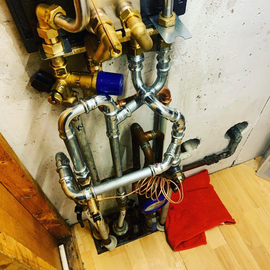 Rørinstallation med metalrør og ventiler på en væg i en kælder, med en rød klud på gulvet.
