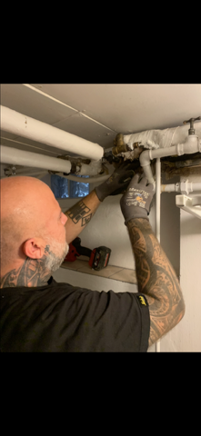 Håndværker arbejder med rørinstallation under et loft, iført handsker og med værktøj i hånden.
