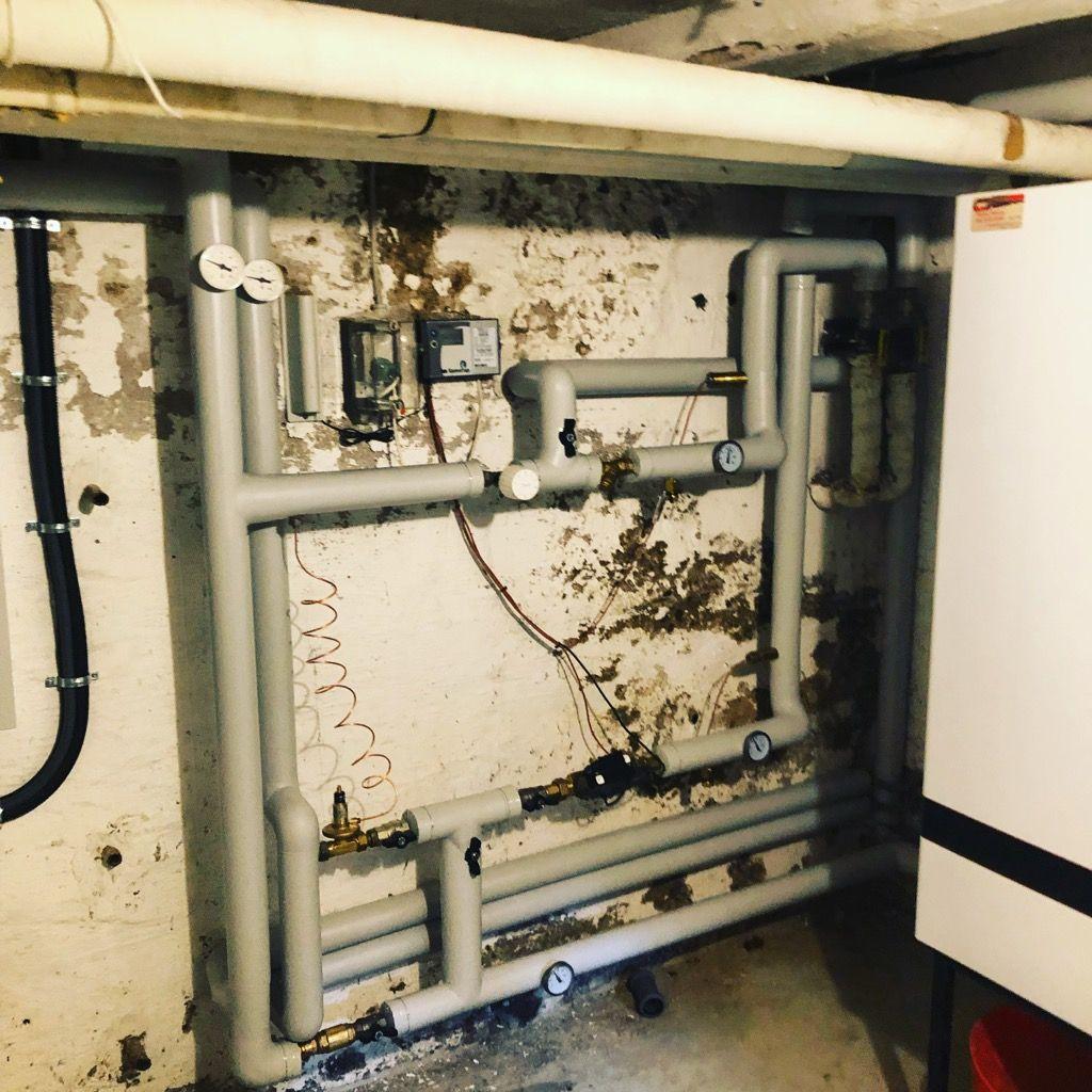 Rørinstallation i kælder med grå rør, målere og synlige skader på væggen.