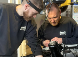 To personer arbejder på en bilmotor, en med pandelampe og den anden med værktøj, begge iført sorte trøjer.