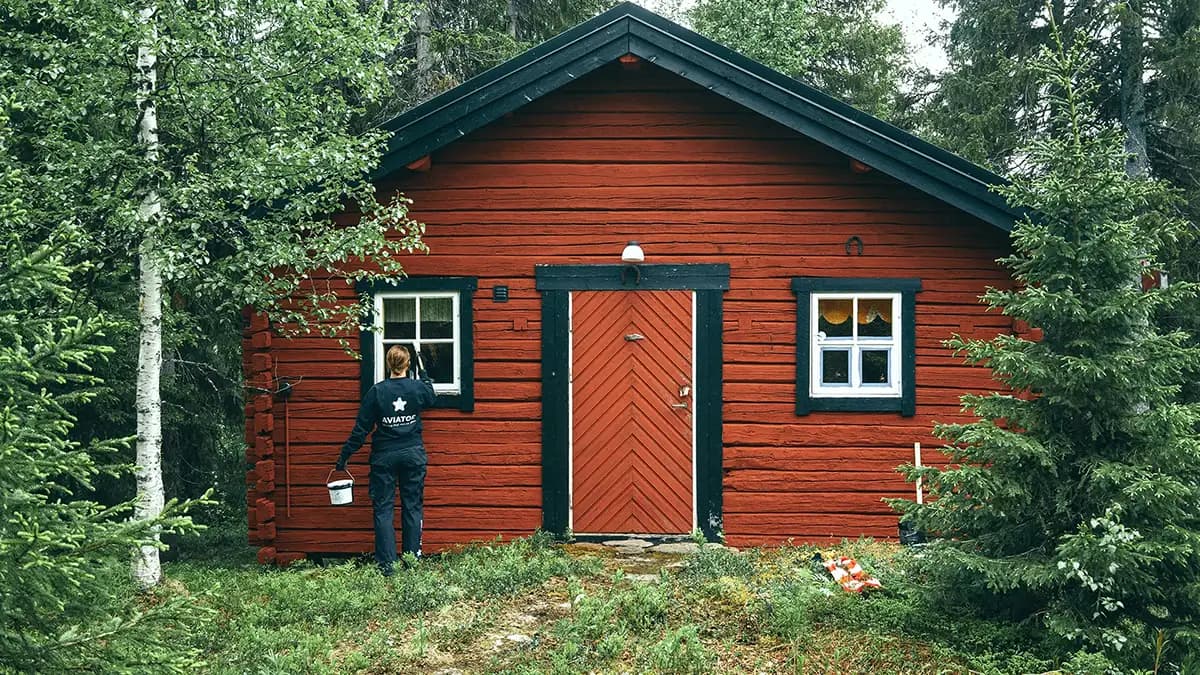 En person der holder en spand står foran et rødt hus omgivet af græs og træer.